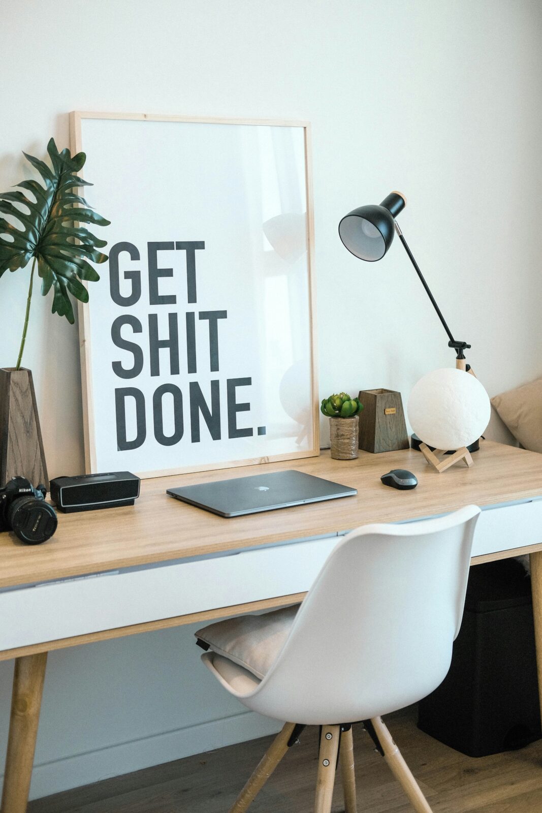 Een minimalistische werkruimte met een houten bureau, een witte stoel, een lamp, een plant en een ingelijste motiverende poster met de tekst "Get Shit Done" vat productiviteit perfect samen. Deze opstelling kan dienen als een inspirerende beschrijving voor iedereen die op zoek is naar focus en creativiteit in hun dagelijkse taken.