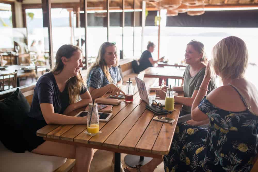 Vier vrouwen zitten rond een houten tafel in een informele binnenomgeving, voeren een gesprek en genieten van een drankje, waarvan één een tablet gebruikt.