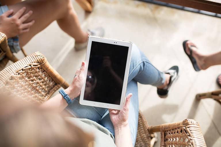 Persoon zittend op een geweven stoel, met een tablet in de hand met een leeg scherm, omringd door anderen in vrijetijdskleding.