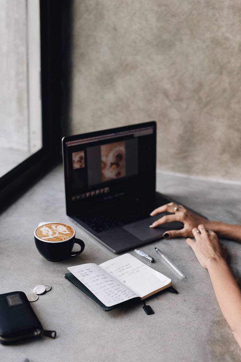 Een persoon die op een laptop werkt, met daarnaast een kop latte, een open notitieboekje met handgeschreven aantekeningen en een paar munten verspreid op een betonnen tafel.