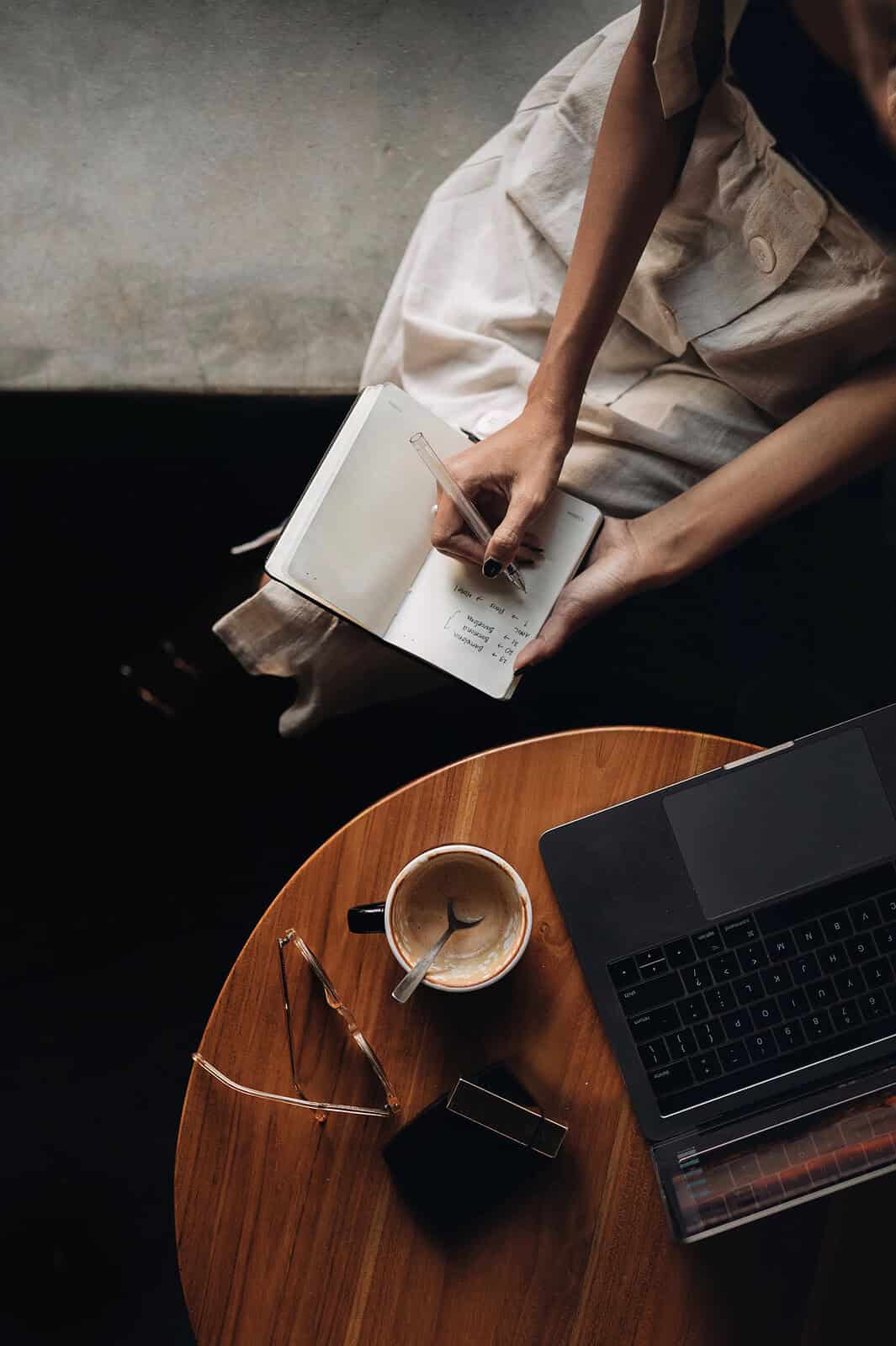 Een persoon zit aan een tafel te schrijven in een notitieboekje, met een kopje koffie, een bril en een laptop in het zicht.