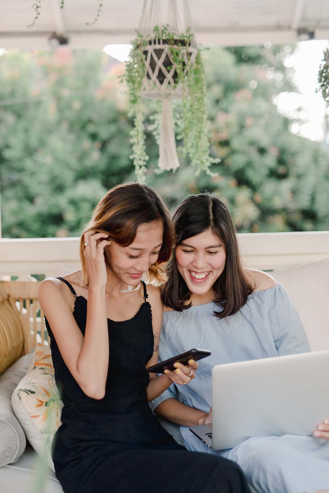 Twee vrouwen zitten buiten op een bank. De een houdt een telefoon vast en lacht, terwijl de ander een laptop gebruikt. Ze lijken allebei te genieten van een moment samen.