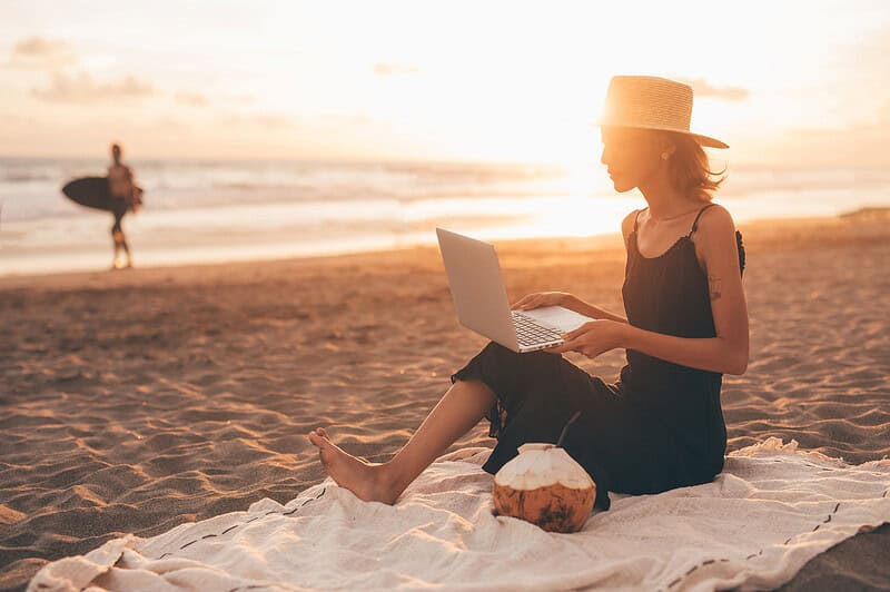 Een persoon zit op het strand met een laptop en een strohoed. Op de achtergrond staan een kokosnoot en een surfer.