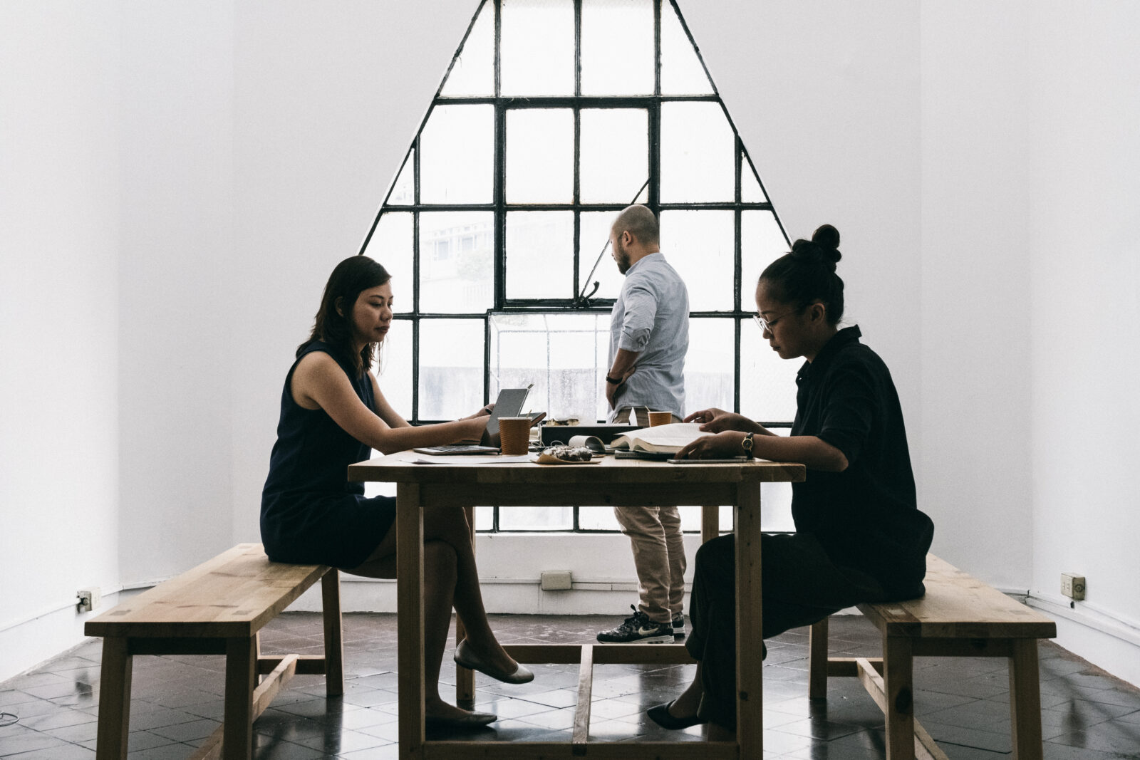 Drie mensen in een minimalistische kamer. Twee zitten aan een tafel en werken op laptops. Ze bespreken automatische concepten, terwijl één persoon nadenkend uit een driehoekig raam staart.