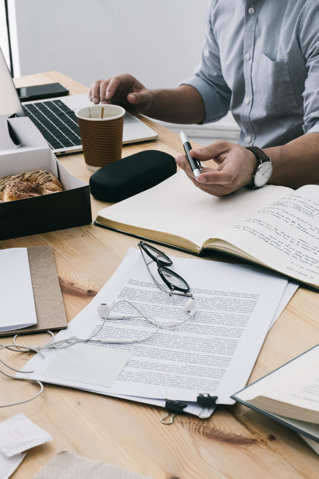 Persoon zit aan een bureau met een laptop, koffie, notitieboekjes, documenten en een bril, en houdt een pen vast.