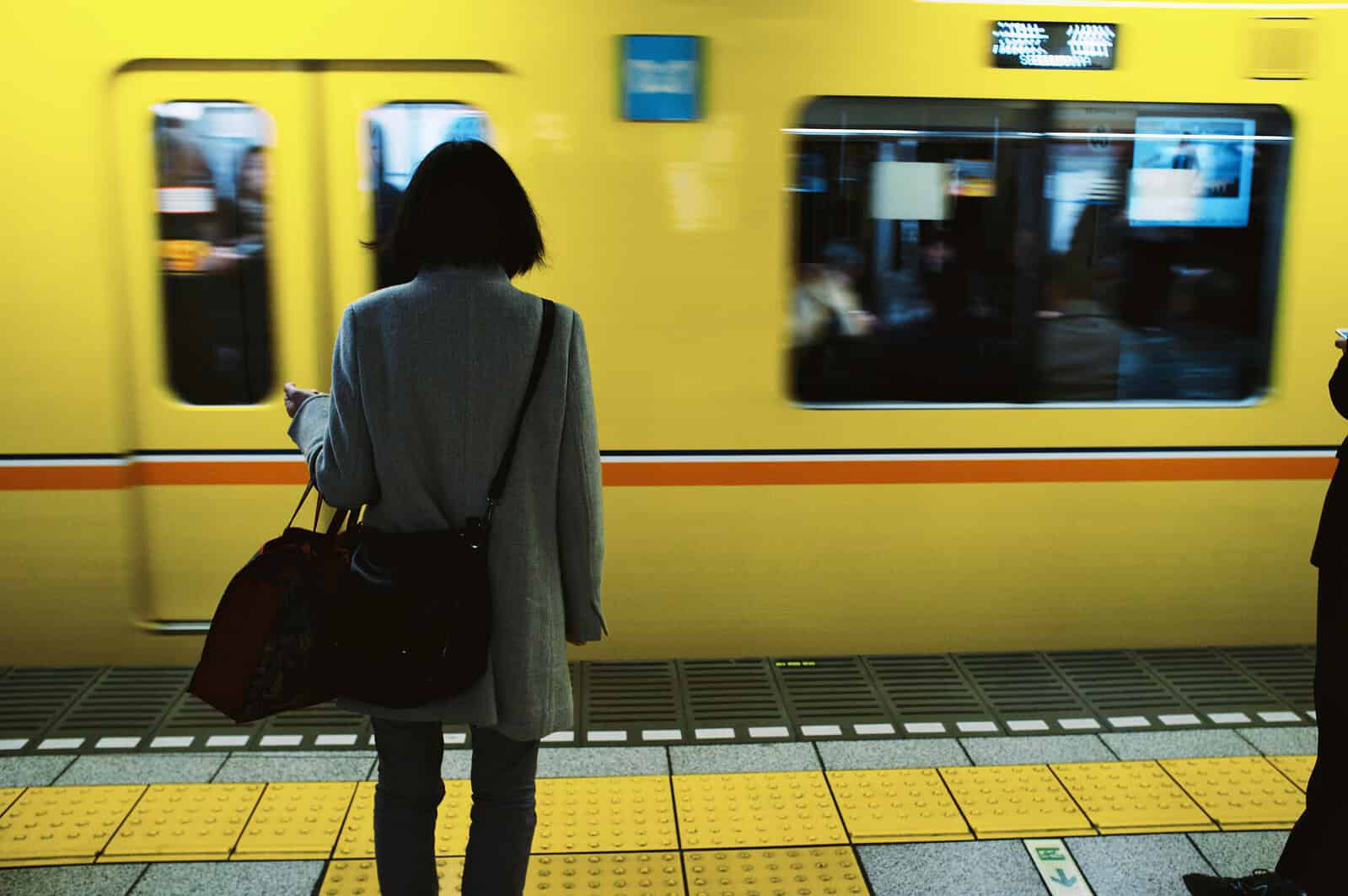 Een persoon met een tas staat op een metroperron terwijl een gele trein voorbijrijdt.