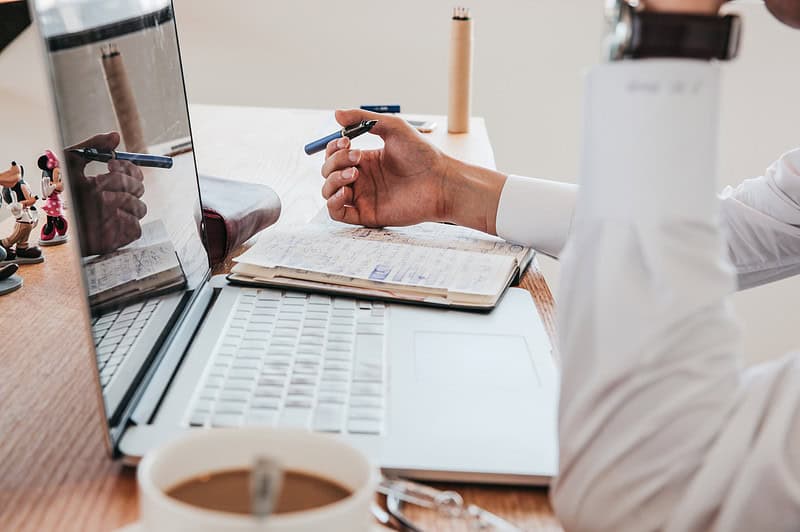 Een persoon in een wit shirt houdt een pen vast terwijl hij een laptop gebruikt. Een notitieboekje, koffiekopje, glazen en beeldjes staan op het bureau.