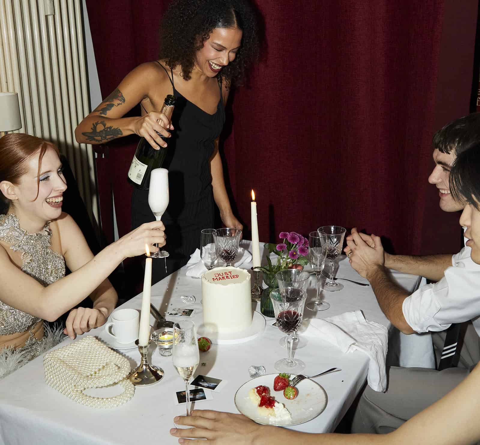 Een vrouw schenkt champagne in aan een tafel met drie mensen. Er staat een taart met "Just Married" op tafel, samen met bloemen, kaarsen en glazen. Ze lijken feest te vieren.