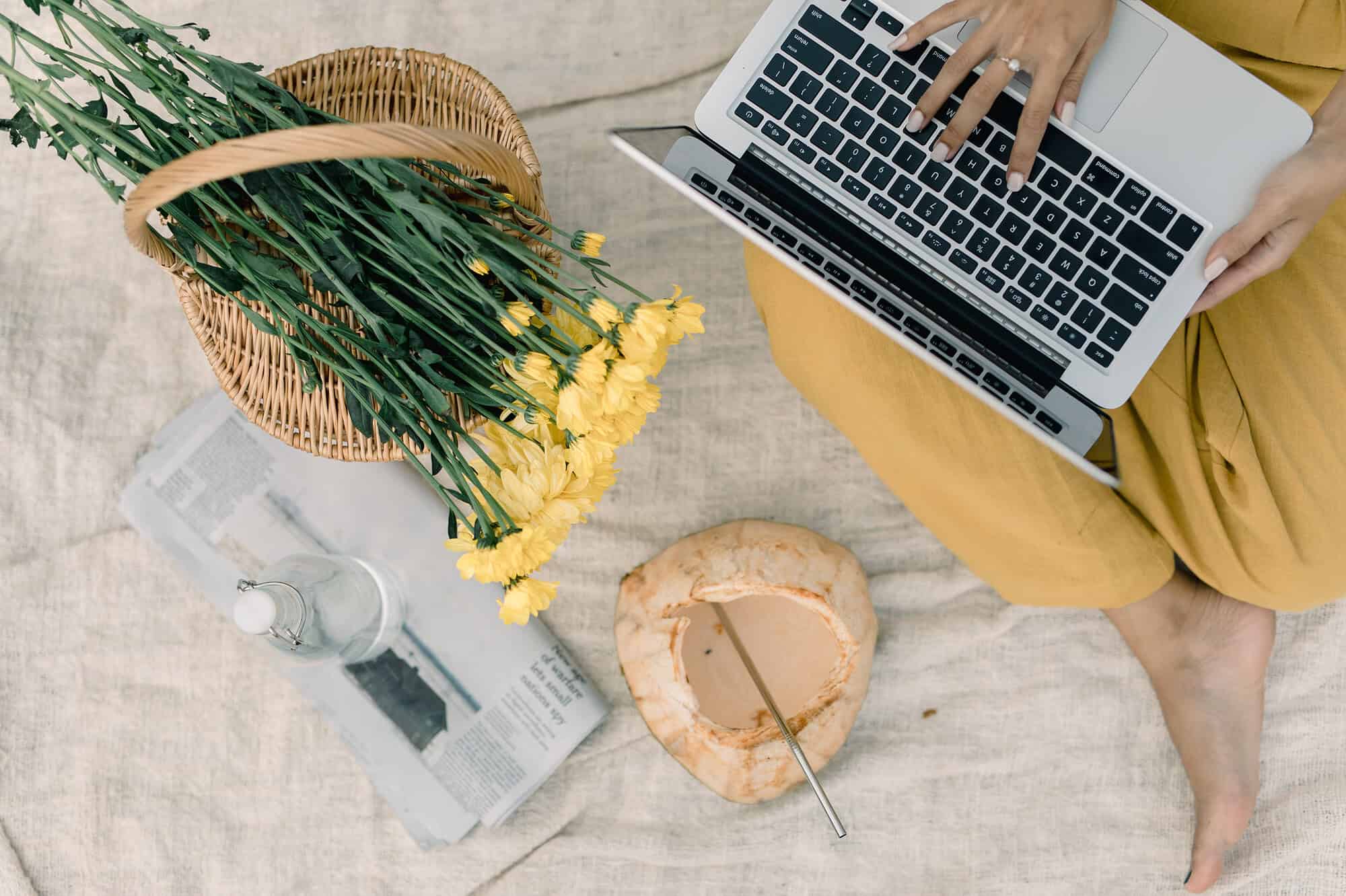 Een persoon in het geel gebruikt een laptop, misschien om te browsen op sociale media of om zijn website te updaten, terwijl hij op een deken zit. In de buurt staan een mand met gele bloemen, een krant, een glazen fles en een kokosnoot met een rietje.