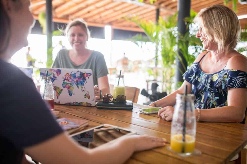 Drie vrouwen zitten aan een houten buitentafel, werken op laptops met kleurrijke hoesjes en drinken drankjes. Ze glimlachen en zijn in gesprek.