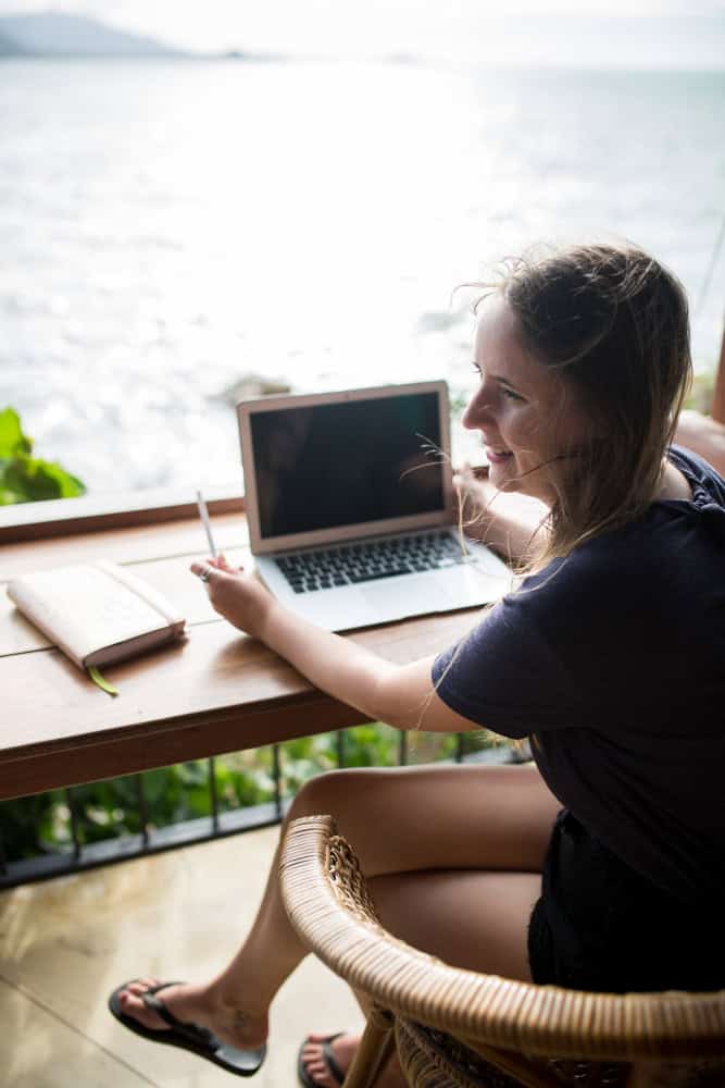 Een persoon die aan een houten tafel zit en op een laptop werkt met een notitieboekje en pen, vlakbij een grote watermassa.