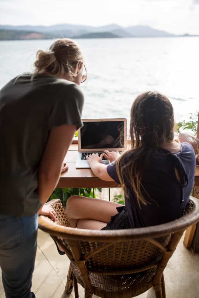 Twee mensen werken buiten op een laptop aan een houten tafel bij het water. De ene persoon zit te typen terwijl de ander naar voren leunt en naar het scherm kijkt.