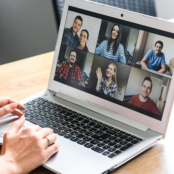 Een persoon neemt deel aan een videoconferentie op een laptop met zes andere mensen op het scherm, geplaatst op een houten tafel naast een kopje koffie.